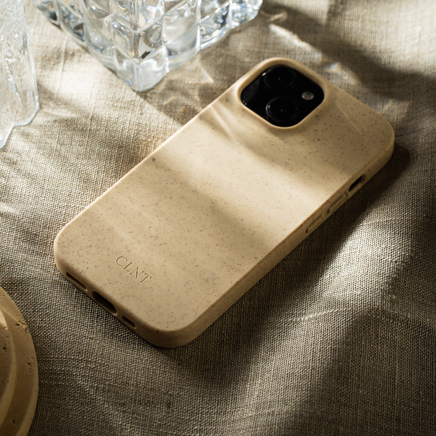 Beige iPhone-deksel fra CLNT – vist liggende på en beige duk med sollys som lyser opp mobildekselet.