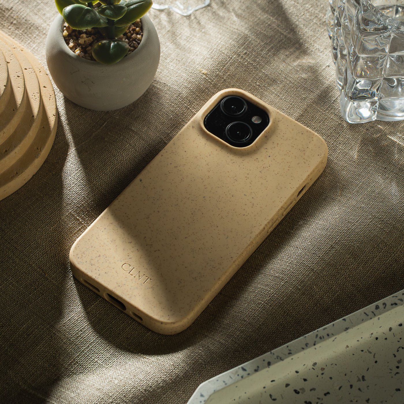 Nærbilde av beige deksel til iPhone fra CLNT - vist liggende på en beige duk med et glass og en liten plante ved siden av samtidig som sollyset kommer inn.