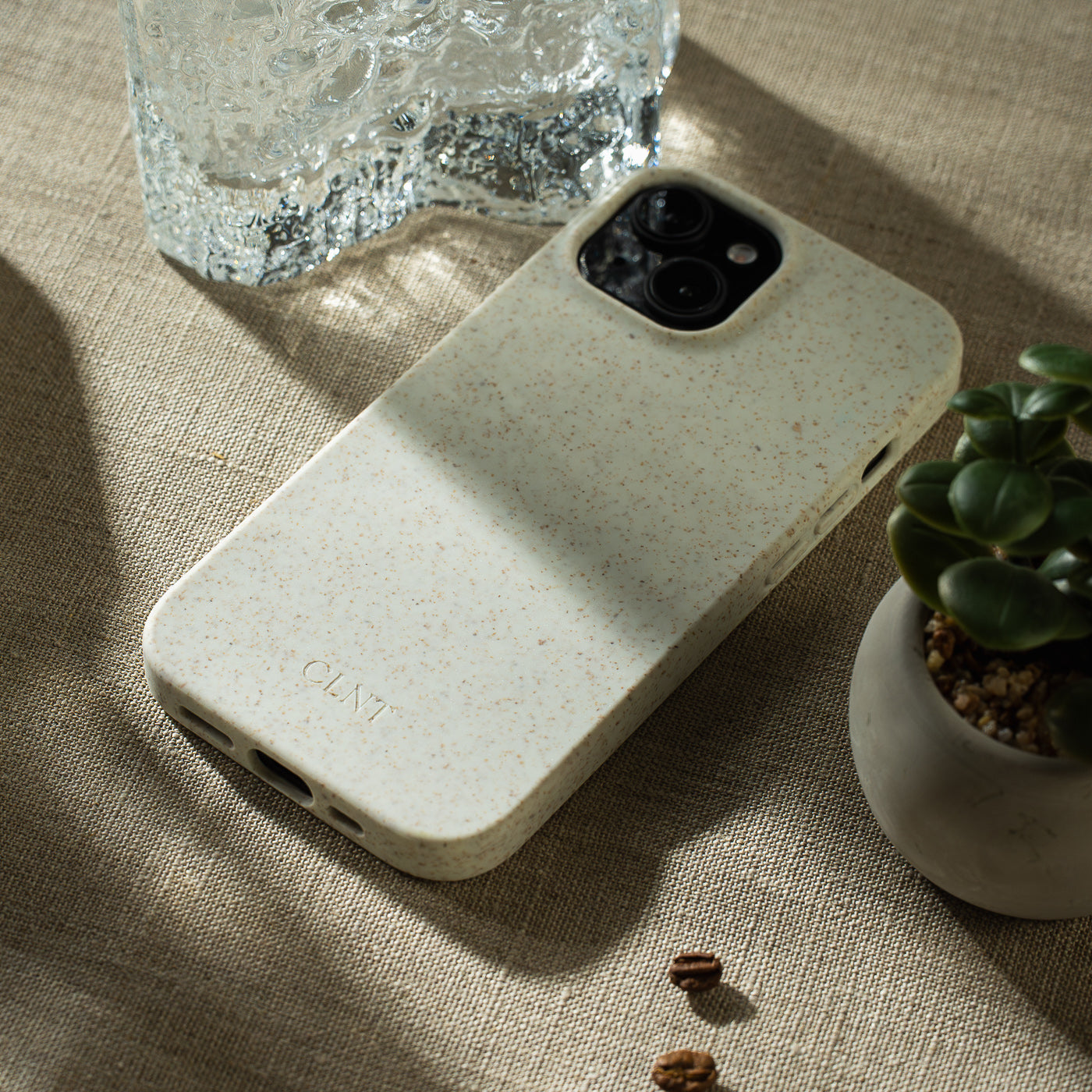 Grått deksel til Iphone fra CLNT - vis på en beige duk med grønn plante og et glass