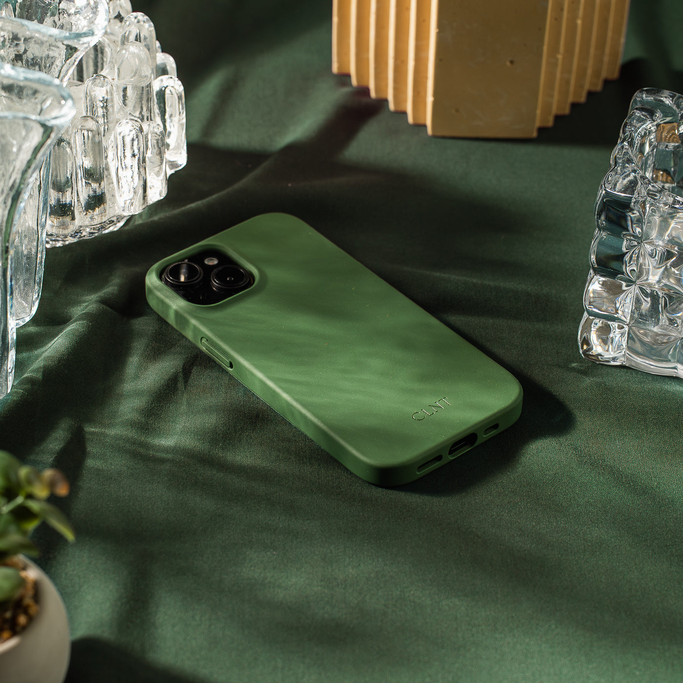 Grønt iPhone-deksel fra CLNT – vist liggende på en grønn duk med glasselementer og en grønn plante rundt.