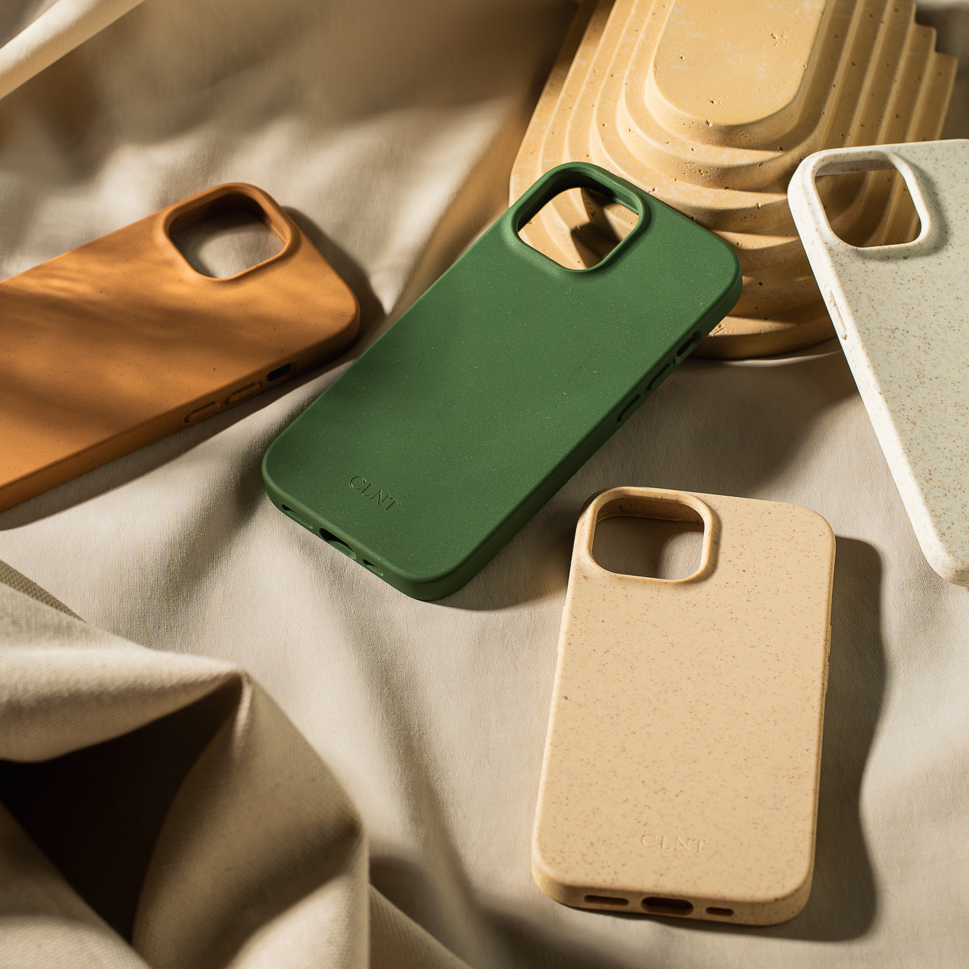 Flere mobildeksler til iPhone fra CLNT som ligger spredt oppå en beige duk med sollys som sprer seg over mobildekslene