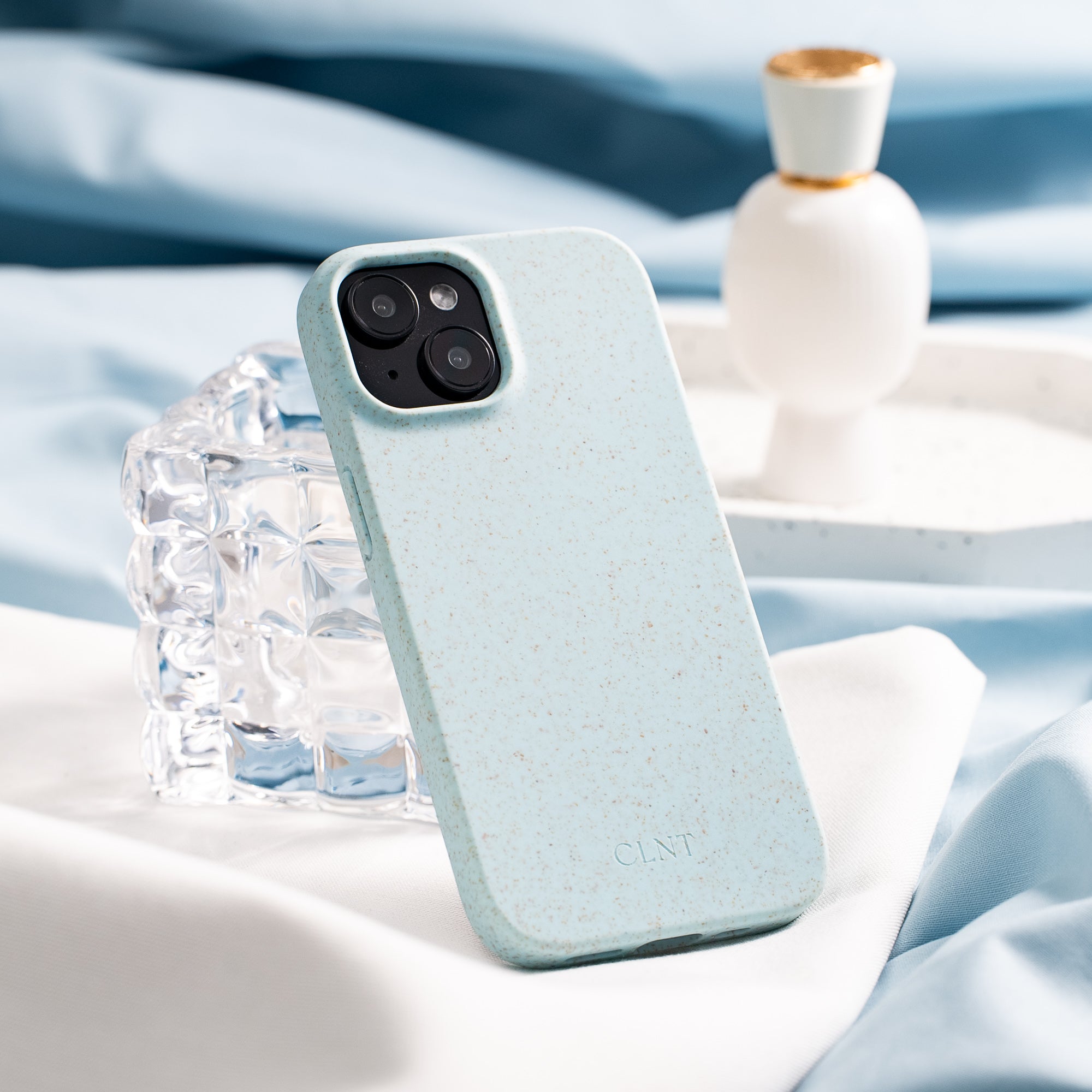 Nærbilde av lyselått deksel til iPhone fra CLNT - vist på en lys duk hvor dekselet er stående inntil et glass.