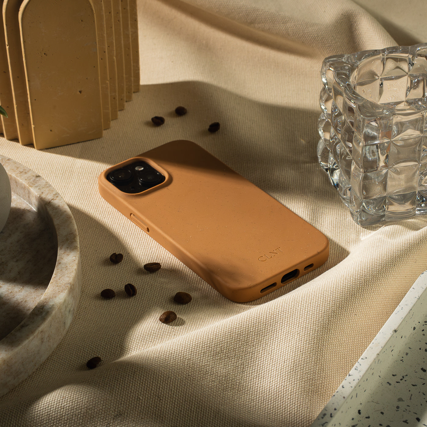 Rustfarget iPhone-deksel fra CLNT – vist liggende på en beige duk omgitt av kaffebønner, marmorfat og glass