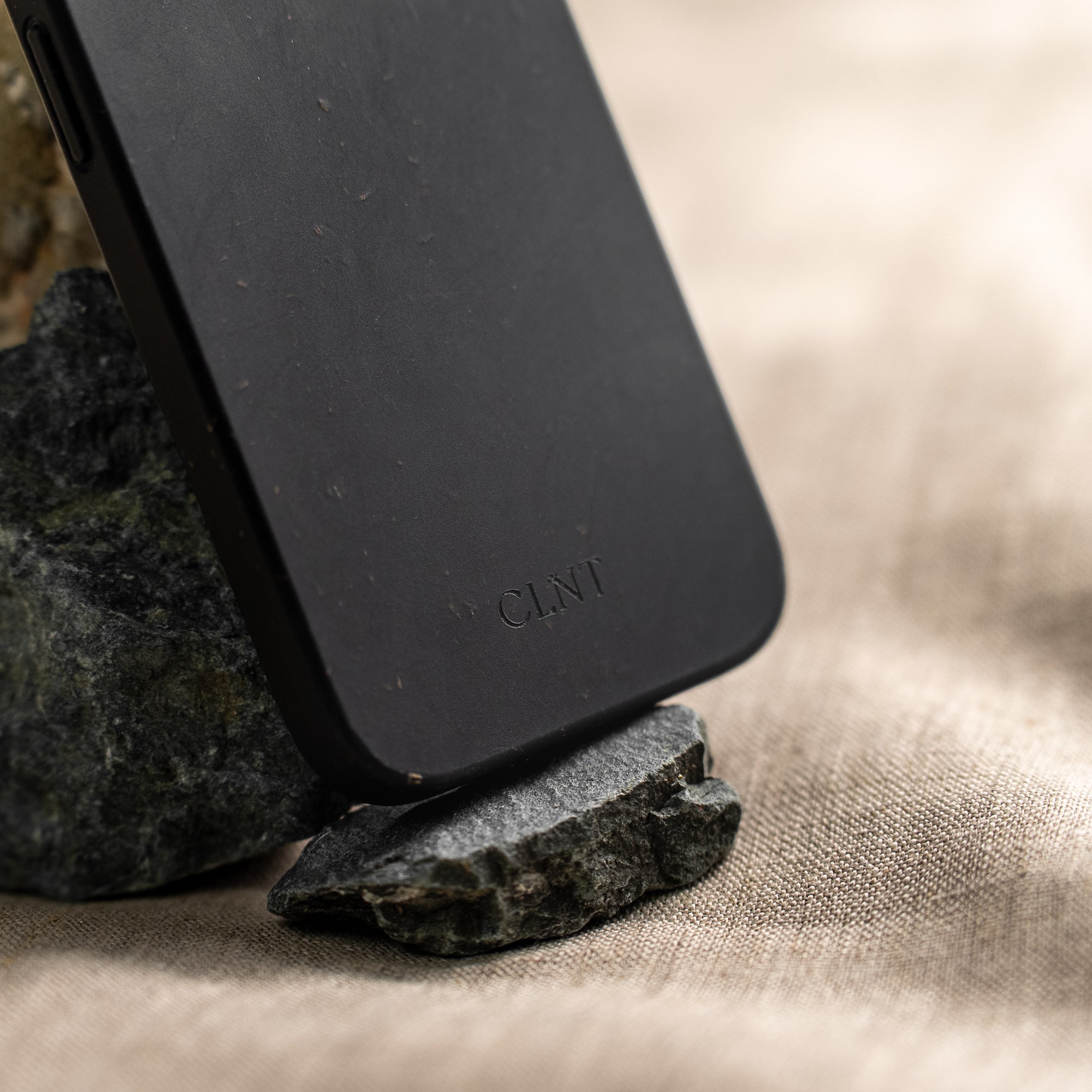 Nærbilde av svart deksel til iPhone fra CLNT - vist stående oppå en mørk stein på en beige duk.