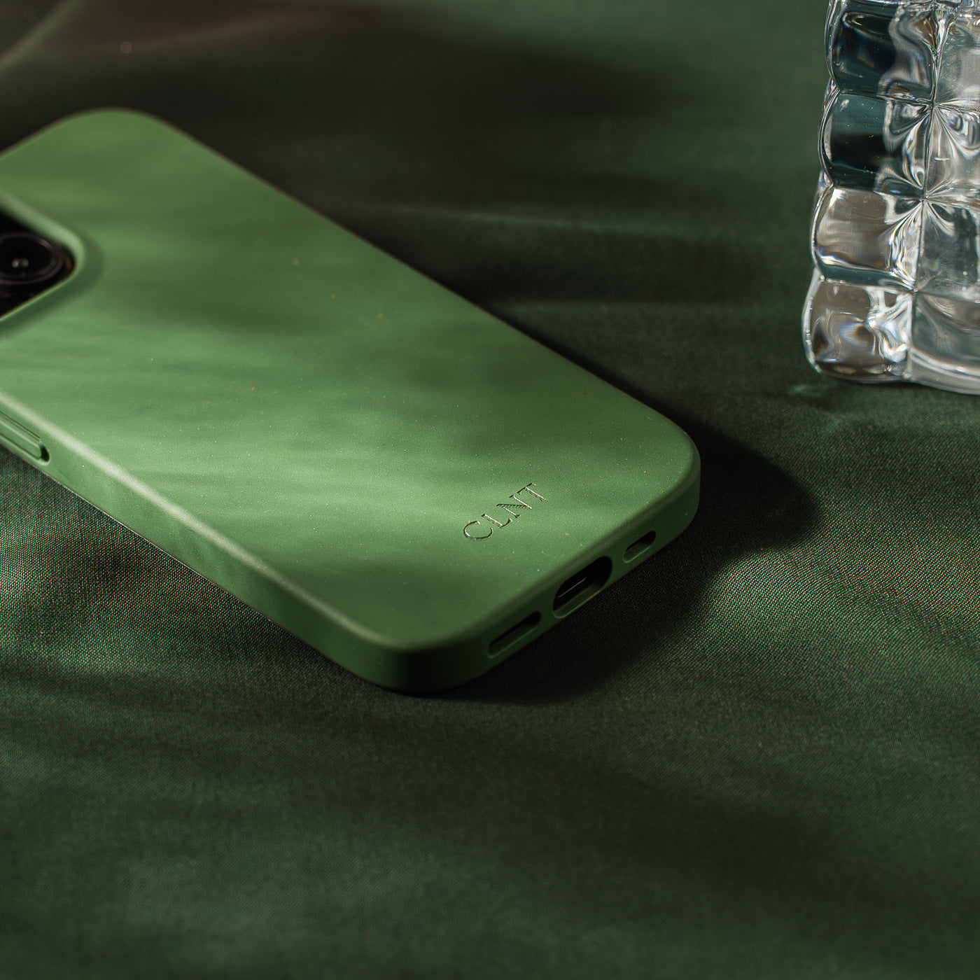Nærbilde av grønt deksel til iPhone fra CLNT - vist liggende på en møkegrønn duk med sollys som kommer inn. 