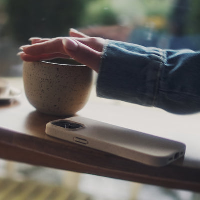 Video av beige deksel til iPhone fra CLNT - vist på en kafè hvor en kvinne drikker kaffe med mobildekselet liggende på bordet. 